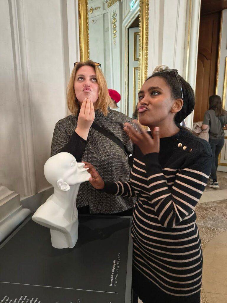 Una viaggiatrice e l'accompagnatrice provano ad imitare l'espressione del curioso busto dell'artista Messerschmidt, che raffigura un uomo con la bocca protesa in avanti, a simulare la forma di un becco.
